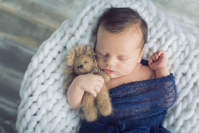 Puede incluir: Un beb&eacute; reci&eacute;n nacido durmiendo pl&aacute;cidamente en una manta de punto blanca, sosteniendo un oso de peluche marr&oacute;n. El beb&eacute; est&aacute; envuelto en una manta azul.