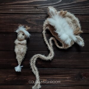 May include: A brown knitted deer hat with white fur trim and a matching knitted deer doll. The hat has long, fluffy strings that can be tied under the chin. The doll has a long, fluffy tail and is standing on a wooden surface.