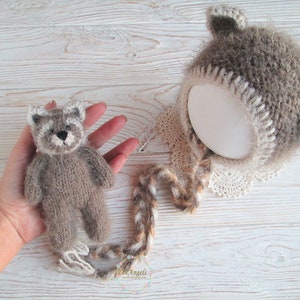 Peut inclure: Un ours en peluche brun tricot&eacute; avec des accents blancs sur le visage, portant un bonnet brun et blanc tricot&eacute; avec une bordure en dentelle blanche. Le bonnet a une longue queue brune et blanche.