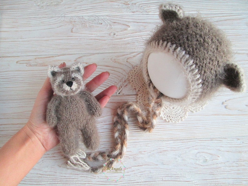 Peut inclure: Un ours en peluche brun tricot&eacute; avec des accents blancs sur le visage, portant un bonnet brun et blanc tricot&eacute; avec une bordure en dentelle blanche. Le bonnet a une longue queue brune et blanche.