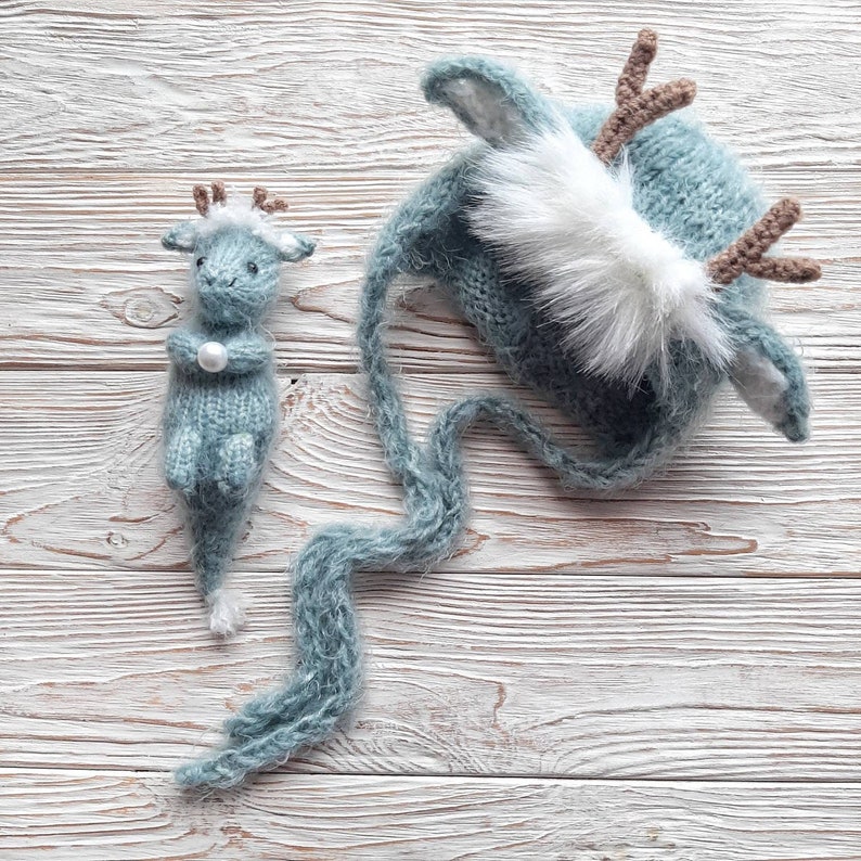 May include: A blue knitted deer hat with a white fur pom-pom and long tail. A smaller blue knitted deer toy is also included.