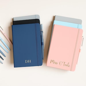 May include: A collection of personalized notebooks in various colors, including navy blue and pink. The navy notebook has the initials "DH" in gold, while the pink notebook is inscribed with "Miss O'Toole". Each notebook has a matching pen.