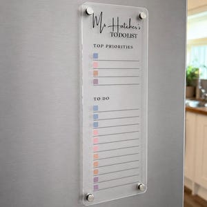 May include: A clear rectangular acrylic dry-erase board with four silver metal standoffs. The board features the printed text Ms. Hatcher's TO DO LIST, with sections for TOP PRIORITIES and TO DO, each containing horizontal lines and colored square icons.