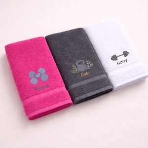 May include: Three personalized gym towels in pink, gray, and white. Each towel features an embroidered fitness-themed design: dumbbells, a kettlebell, and a barbell. Names are embroidered below the designs.
