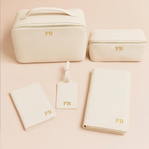 Peut inclure: Ensemble d'accessoires de voyage crème, comprenant une trousse de maquillage, un petit étui, un porte-passeport, une étiquette de bagage et un portefeuille zippé. Chaque article a des fermetures éclair dorées et les initiales "PB" en lettres dorées.
