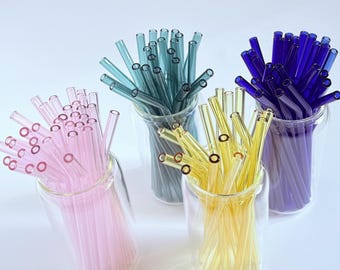 May include: A selection of reusable glass straws in shades of pink, teal, yellow, and blue. Each set of straws is presented in a clear glass cup. The straws are straight with a circular opening.