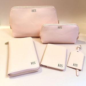 Peut inclure: Ensemble de cinq accessoires rose pâle assortis : deux trousses de maquillage zippées, un portefeuille, un porte-passeport et une étiquette de bagage. Chaque article porte les initiales "HS" en noir. Les sacs ont des fermetures éclair dorées.