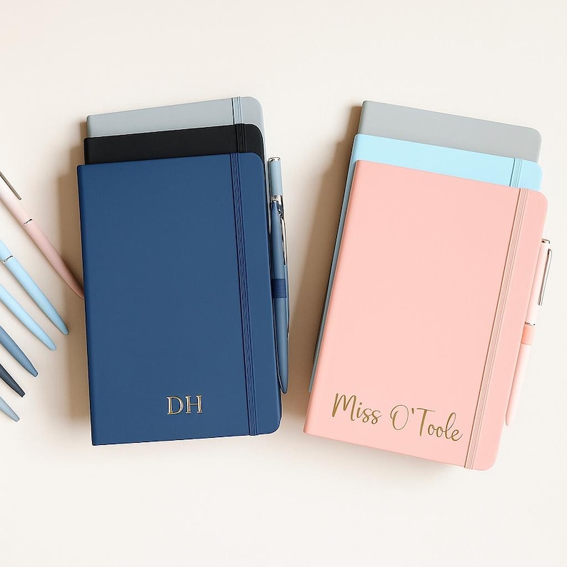 May include: Two stacks of hardcover notebooks with elastic closures and matching pens. The left stack features a navy blue notebook with gold DH initials. The right stack features a pink notebook with gold Miss O'Toole text, plus blue and gray notebooks.