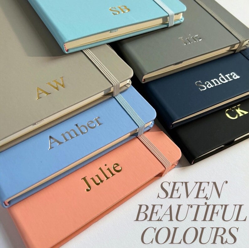 Puede incluir: Una colecci&oacute;n de siete cuadernos personalizados en varios colores, como azul claro, gris y coral. Cada cuaderno presenta un nombre o iniciales en dorado o plateado, con cierre de banda el&aacute;stica. Tambi&eacute;n se ve el texto "SEVEN BEAUTIFUL COLOURS".