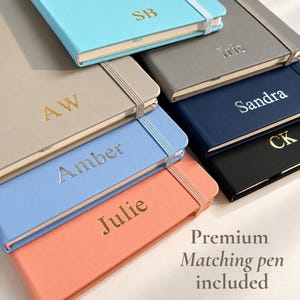 May include: A collection of rectangular notebooks in various colours including light blue, beige, periwinkle, coral, grey, navy, and black. Each notebook features an elastic closure band and personalised gold or silver foil text like SB, AW, Amber, Julie, Iris, Sandra, and CK.