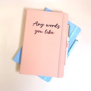 May include: A collection of notebooks in pastel shades. The top notebook is pale pink with the text "Any words you like" in black script. A pale pink pen rests on the side. A light blue notebook is visible.