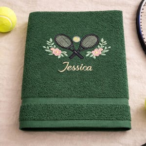 May include: A dark green bath towel featuring an embroidered tennis design. The design includes crossed tennis rackets, a tennis ball, and floral accents. The name "Jessica" is embroidered below the design.