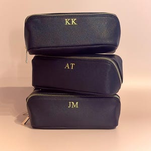 May include: Three navy blue zippered pencil cases stacked on top of each other. Each case has gold-colored initials: KK, AT, and JM. The cases have a rectangular shape with rounded edges and a gold zipper.