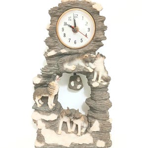 May include: A decorative clock featuring a wolf theme. The clock face has a gold-colored bezel and black numbers. The clock is surrounded by a gray, rocky structure with several wolf figurines. The wolves are in various poses, including howling and interacting.