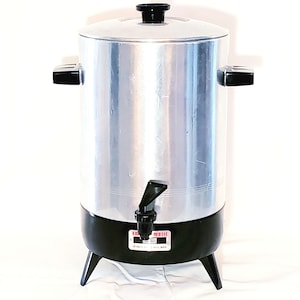 May include: A vintage, silver-toned electric coffee urn with a black base and lid. The urn has a spigot, two black handles, and a black knob on top. The brand name "E-Z-MATIC" is visible on the front. It stands on three black legs.