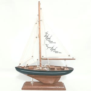 May include: A wooden sailboat model with a dark green hull and a light brown deck. The sails are white with black text that reads "The will of God will never lead you where the grace of God cannot keep you."