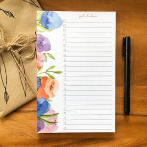 May include: A white notepad with a floral watercolor design in blue, purple, and orange. The notepad has a checklist format with the words "get it done" in a light orange script. A black pen rests on the wooden surface.