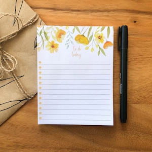 May include: A white notepad with a floral design featuring yellow flowers and green leaves. The notepad has a lined interior with a dotted checklist and the text "To do today" at the top. A black pen is lying next to the notepad.