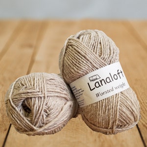 Puede incluir: Dos ovillos de lana beige de peso grueso, uno con una etiqueta que dice "Lanaloft Worsted weight".