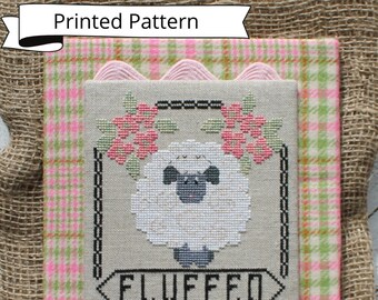 May include: A cross-stitch pattern featuring a white sheep with black eyes and nose, surrounded by pink flowers and a sign that reads "FLUFFED". The pattern is framed with a pink and green plaid border.