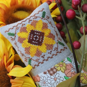 May include: A small, grey linen cushion with a cross-stitch design of a sunflower in shades of yellow, brown, and orange. The cushion is trimmed with a white fabric with a floral pattern.