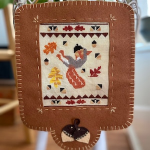 May include: A brown felt wall hanging with a cross-stitched autumn scene. The scene features a figure with falling leaves and acorns. The felt is stitched around the edges with a lighter thread. Two felt acorns are attached at the bottom.