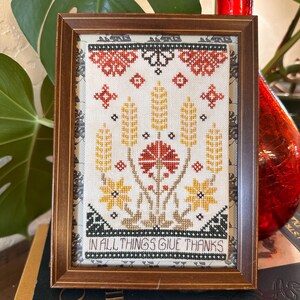 May include: Framed cross-stitch artwork with a floral and wheat design in red, yellow, and brown. The text "IN ALL THINGS GIVE THANKS" is in black. The frame is made of dark brown wood. The artwork is a decorative piece.