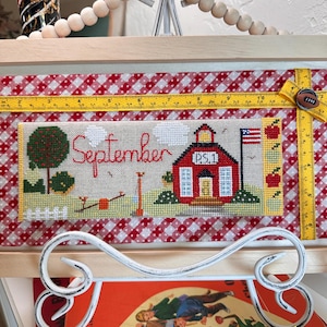 May include: Cross-stitch artwork featuring a red schoolhouse with the text "September" and "P.S. 1". The artwork includes a tree, a seesaw, and an American flag. A yellow measuring tape is attached to the frame, which is set against a red and white checkered background.