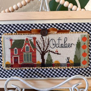 May include: Cross-stitch artwork featuring a red house with a green roof, a bare tree, and the word "October." The design includes pumpkins, a black cat, and a decorative border. The artwork is framed in a wooden frame.