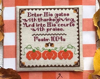 Könnte beinhalten: Kreuzstich-Stickerei mit beigefarbenem Hintergrund, braunem Rand und einem Vers aus Psalm 100,4a: "Geht durch seine Tore mit Dank, in seine Vorhöfe mit Lobgesang." Der Vers ist von einer Reihe orangefarbener Kürbisse mit grünen Ranken umgeben.
