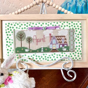 Puede incluir: Un diseño de punto de cruz enmarcado que presenta una casa con un techo morado y la palabra "Abril" en letras rosas. La casa está rodeada de árboles y conejos blancos. El marco tiene un fondo de lunares verdes.