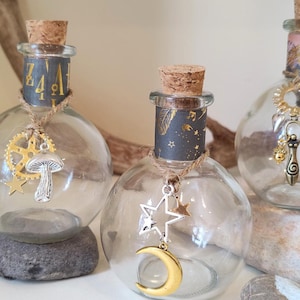 May include: Four clear glass bottles with cork stoppers and decorative charms hanging from them. The charms include a crescent moon, a mushroom, a star, and a wand. The bottles are sitting on small rocks.