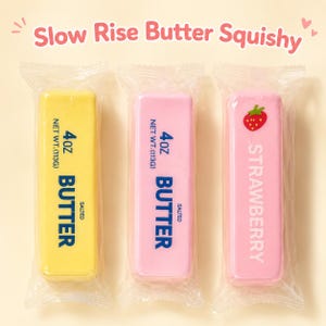 Puede incluir: Tres juguetes squishy rectangulares de subida lenta en envoltorios de plástico transparente. Uno es amarillo con la etiqueta 113g BUTTER SALTED, otro rosa claro con 113g BUTTER SALTED y otro rosa claro con un gráfico de fresa y STRAWBERRY.