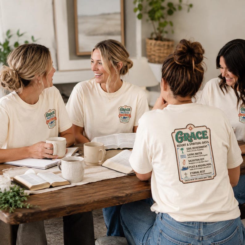 Puede incluir: Cuatro mujeres con camisetas color crema con el logo "Grace Motel" y texto en la parte delantera y trasera. La parte trasera de la camiseta presenta una lista de conceptos espirituales. Est&aacute;n sentadas alrededor de una mesa de madera con libros abiertos y tazas.