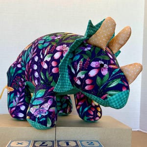 May include: A stuffed triceratops toy made of dark purple fabric with a colorful floral and butterfly print, featuring three light orange polka-dot horns. Four wooden alphabet blocks are arranged in front, spelling the word DINO in capital letters.