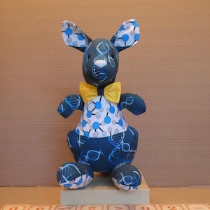 May include: A blue and white kangaroo plush toy wearing a yellow bow tie sits on top of a stack of wooden blocks that spell out "kangaroo".