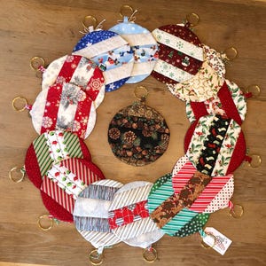 May include: A collection of 14 handmade Christmas ornaments, each featuring a different fabric pattern with red, white, green, and blue colors. The ornaments are round and have a gold ring attached to the top. The ornaments are arranged in a circle on a wooden surface. The text "Crook's Nook Crafts" is visible on a tag.