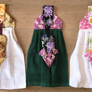May include: Three kitchen towels with a fox design. The towels are white with a patterned fabric loop at the top. The fox is made of fabric and hangs from the towel. The first towel has a brown and gold patterned loop and fox. The second towel has a green towel with a black and purple patterned loop and fox. The third towel has a pink and white patterned loop and fox.