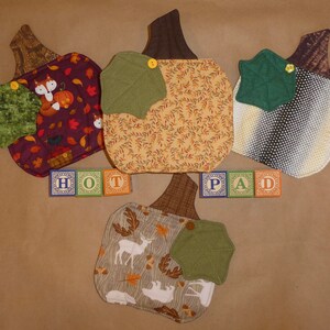 May include: Four quilted pumpkin-shaped hot pads with different fabric patterns. The hot pads are arranged in a row with the words "HOT PAD" spelled out in colorful blocks.