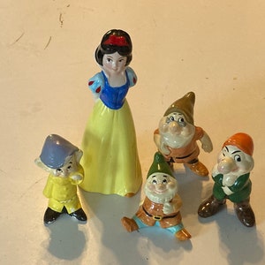 May include: A set of Snow White and the Seven Dwarfs figurines. Snow White is wearing a yellow dress with a blue bodice and a red bow in her hair. The dwarfs are wearing colorful clothing and have different facial expressions.