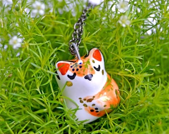Pode incluir: Um colar com um pingente de cerâmica branca com um design de gato laranja e preto pintado à mão. O gato está sentado com as patas enfiadas debaixo do corpo. O colar está em uma corrente prateada e está descansando sobre um leito de grama verde.