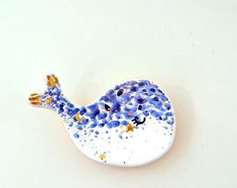 May include: A white ceramic whale dish with blue and gold accents. The whale has a whimsical design with a crescent moon and a star on its body.
