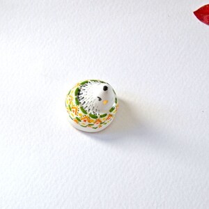 May include: A small, ceramic hedgehog figurine. The hedgehog is white with a black nose and eyes. Its back is decorated with a pattern of green, yellow, and orange shapes. The figurine is on a white surface.