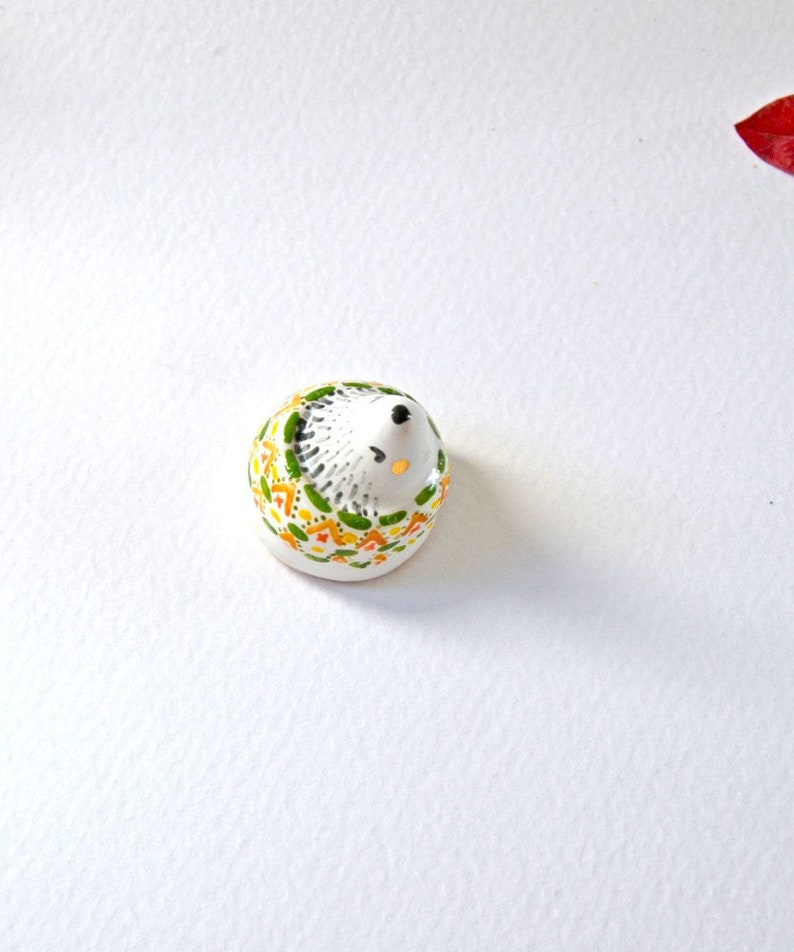 May include: A small, ceramic hedgehog figurine. The hedgehog is white with a black nose and eyes. Its back is decorated with a pattern of green, yellow, and orange shapes. The figurine is on a white surface.