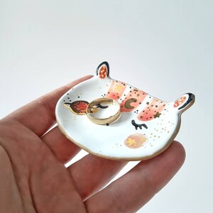 Op de afbeelding: Een witte keramische ringenschotel met een roze en gouden ontwerp van een slapende kat met sterren en een halve maan.