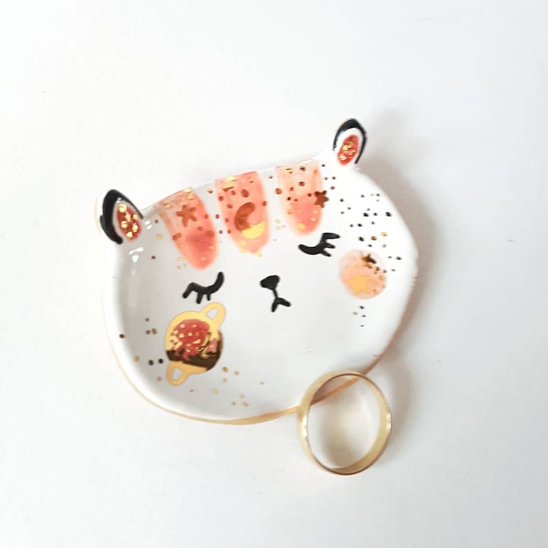 Op de afbeelding: Witte keramische ringenschotel in de vorm van een kattenkop met roze en gouden accenten. De schotel heeft een gouden ring erin.