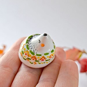 May include: A small, ceramic hedgehog figurine. The hedgehog has a white body with a black and gray back, a black nose, and orange accents. The figurine is decorated with green, orange, and yellow floral patterns around the base.