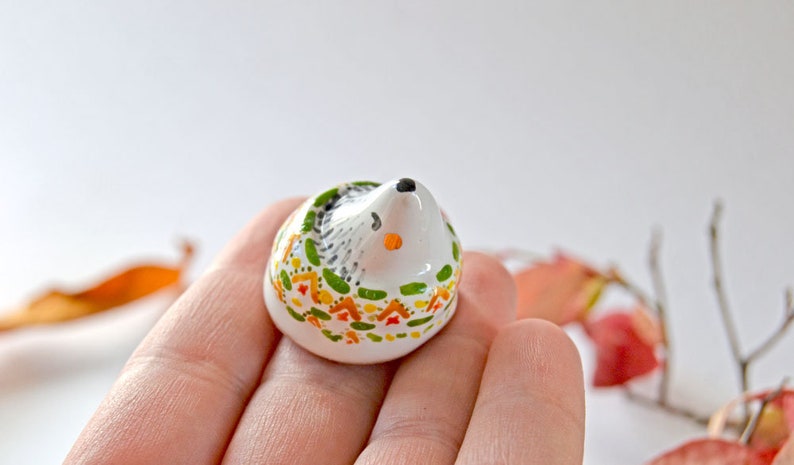 May include: A small, ceramic hedgehog figurine. The hedgehog has a white body with a black and gray back, a black nose, and orange accents. The figurine is decorated with green, orange, and yellow floral patterns around the base.
