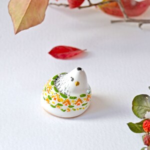 May include: A small, ceramic hedgehog figurine with a white body and a decorative pattern of green, orange, and yellow accents. The hedgehog has a black nose and eyes, and is adorned with small, sparkling embellishments. The figurine is set against a white background.