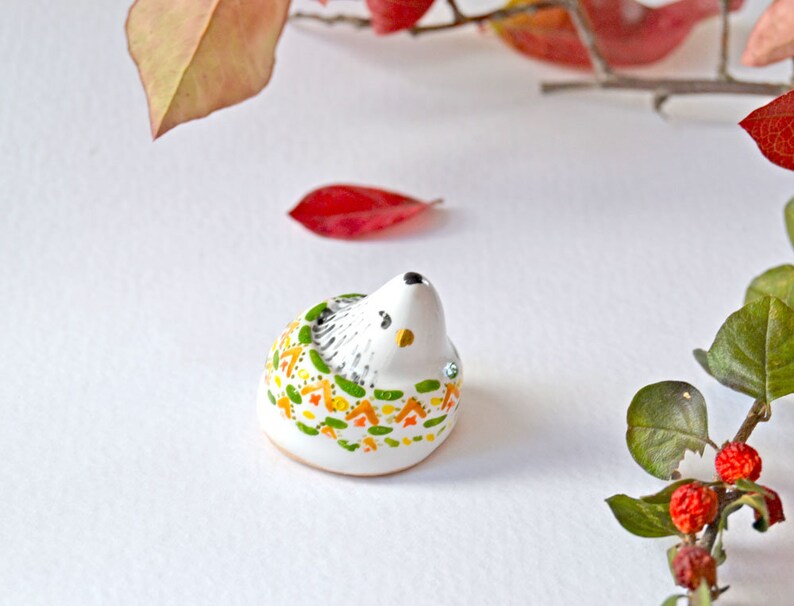 May include: A small, ceramic hedgehog figurine with a white body and a decorative pattern of green, orange, and yellow accents. The hedgehog has a black nose and eyes, and is adorned with small, sparkling embellishments. The figurine is set against a white background.